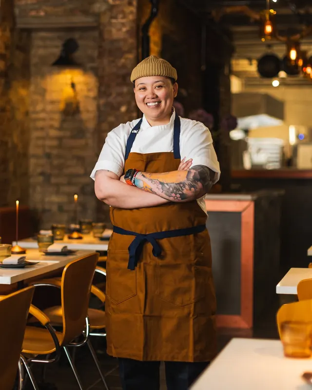 Chef Frances Tariga, arms crossed, in a white chef's jacket and ochre apron